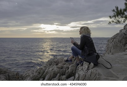 Beautiful blonde girl is seating on the stones near of the sea with tablet in hands and watching into the distance. 