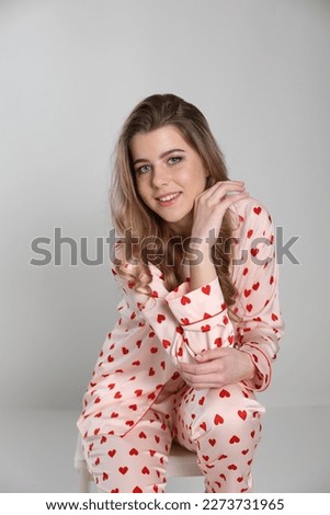 beautiful blonde girl sits on a chair on a white background in pajamas. pajamas with hearts. Valentine's Day. studio photo shoot in satin pajamas with a heart print. clothes for sleep and home