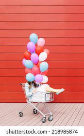 Beautiful blonde girl sitting in shopping cart, holding color balloons in both hands hiding her face. Sunny day, outside Stock Photo