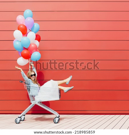 Beautiful blonde girl sitting in shopping cart, color balloons in her one hand. Smiling, laughing. Sunny day, outside