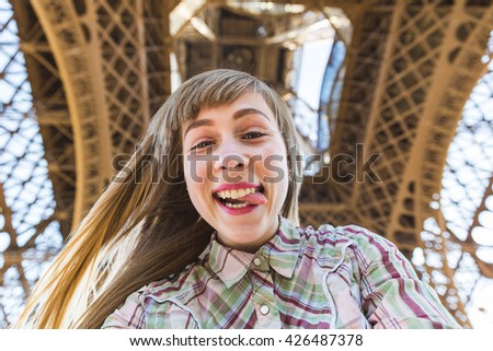 Beautiful blonde girl taking a selfie under the Eiffel Tower in Paris. She is looking at camera, smiling and grimacing with the tongue out of the mouth. Travel and lifestyle concepts.
