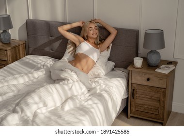 A beautiful blonde girl in a white top wakes up in the morning in a cozy bed. The concept of a beautiful morning, wake up with a good mood