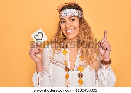 Beautiful blonde hippie woman with blue eyes wearing sunglasses holding reminder with heart surprised with an idea or question pointing finger with happy face, number one