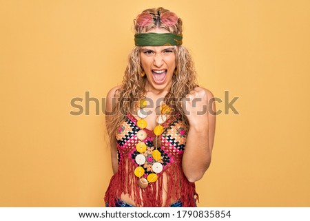 Beautiful blonde hippie woman wearing sunglasses and accessories over yellow background angry and mad raising fist frustrated and furious while shouting with anger. Rage and aggressive concept.