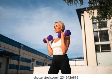 Beautiful blonde middle age woman doing weights exercises with dumbbells outdoors, sunny summer evening. Healthy active lifestyle, body conscious  - Powered by Shutterstock