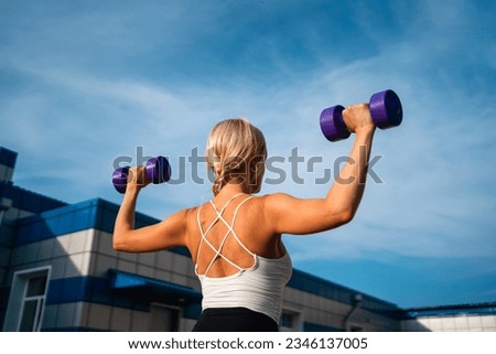 Beautiful blonde middle age woman doing weights exercises with dumbbells outdoors, sunny summer evening. Healthy active lifestyle, body conscious 