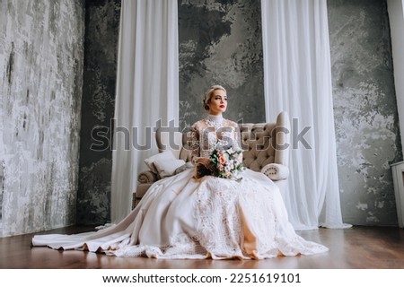 A beautiful blonde model bride in a long lace dress sits on a sofa in the studio, indoors with a bouquet of flowers in her hands on a gray background. Wedding photography, portrait.