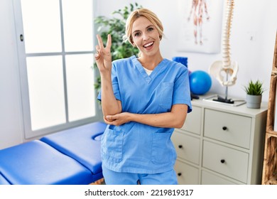 Beautiful Blonde Physiotherapist Woman Working At Pain Recovery Clinic Smiling With Happy Face Winking At The Camera Doing Victory Sign. Number Two. 