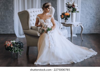 A beautiful blonde princess bride in a white lace dress with a crown, a diadem sits on a sofa in the studio, indoors with a bouquet in her hands. Wedding photography, close-up portrait. - Powered by Shutterstock