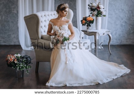 A beautiful blonde princess bride in a white lace dress with a crown, a diadem sits on a sofa in the studio, indoors with a bouquet in her hands. Wedding photography, close-up portrait.