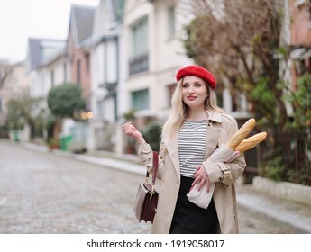 A beautiful blonde in a red beret, beige raincoat and a striped blouse holds French baguettes in her hands. Classic French girl in Paris. - Shutterstock ID 1919058017