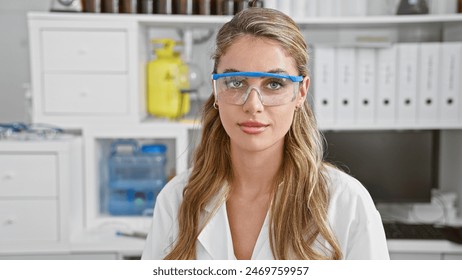 Beautiful blonde scientist expressing seriousness in safety glasses while engaged in promising medical research within the confines of her modern laboratory workspace - Powered by Shutterstock