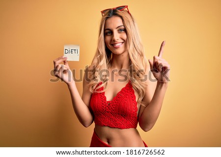 Beautiful blonde slim woman on vacation wearing bikini holding reminder with diet message surprised with an idea or question pointing finger with happy face, number one