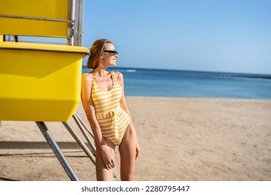 Beautiful blonde smiling woman standing in the sun at the beach, wearing fashionable one piece swimwear. Blue and yellow colors. Traveler. Tourism. Copy space.  