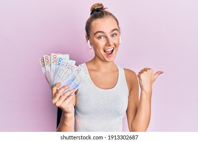 Beautiful blonde sport woman holding 20 swedish krona banknotes pointing thumb up to the side smiling happy with open mouth 