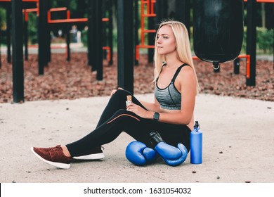 Beautiful Blonde Sports Girl Eating Muesli And Cereal Snack While Sitting On Sports Ground. Attractive Young Fitness Woman Posing With Protein Bar In Hand While Resting After Boxing Training Outdoors