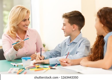 Beautiful blonde teacher sitting at desk, wearing in pink shirt, holding blue pencil in her hand and telling her little pupils technique of drawing. Children listening carefully to her and smiling.