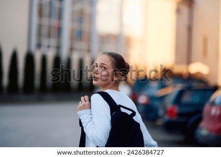 A beautiful blonde teenager returns home from school during the sunset, carrying her school backpack, in a serene and peaceful urban setting