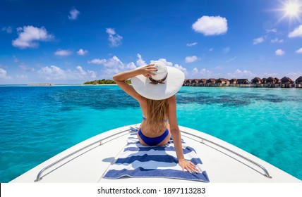 A beautiful, blonde woman in bikini with hat sits on a yacht and enjoys the summer holiday over the tropical ocean of the Maldives islands