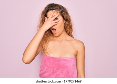 Beautiful Blonde Woman With Blue Eyes Wearing Towel Shower After Bath Over Pink Background Peeking In Shock Covering Face And Eyes With Hand, Looking Through Fingers With Embarrassed Expression.