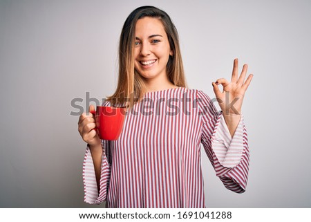 Beautiful blonde woman with blue eyes drinking red mug of coffee over white background doing ok sign with fingers, excellent symbol