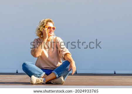 Beautiful blonde woman in casual clothes sitting and talking on the phone. Happy beautiful blonde caucasian woman in glasses and fashionable clothes on a light background. 