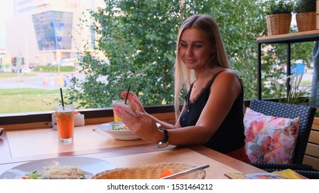 Beautiful Blonde Woman Chatting On The Phone, Talking On Skype And Drinking A Cocktail, Sitting In A Cafe On The Street