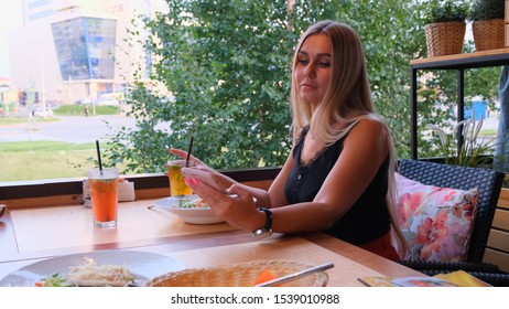 Beautiful Blonde Woman Chatting On The Phone, Talking On Skype And Drinking A Cocktail, Sitting In A Cafe On The Street