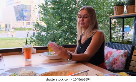 Beautiful Blonde Woman Chatting On The Phone, Talking On Skype And Drinking A Cocktail, Sitting In A Cafe On The Street