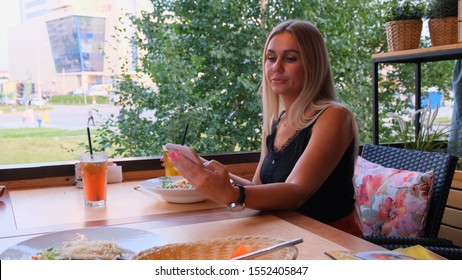 Beautiful Blonde Woman Chatting On The Phone, Talking On Skype And Drinking A Cocktail, Sitting In A Cafe On The Street