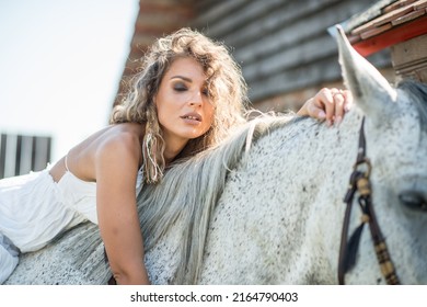 Beautiful blonde woman with curly hair and horse. Portrait of a girl with white dress and her horse. Beautiful girl interacting and having fun with a horse at the ranch