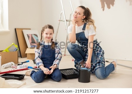 Beautiful blonde woman with cute 8-year-old daughter are sitting on floor in middle of a room undergoing renovation. near them are many boxes of small things as well as cans of paints and rollers.