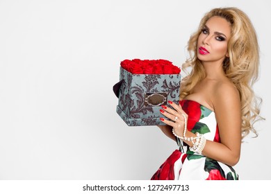 Beautiful blonde woman in an elegant evening gown with red roses, holding a Valentine's gift, a flowerbox with flowers. Beauty model posing in the studio on a white background.