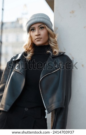 beautiful blonde woman in fashion clothes with a knitted hat and a black stylish sweater with a leather jacket stands near a wall in the city