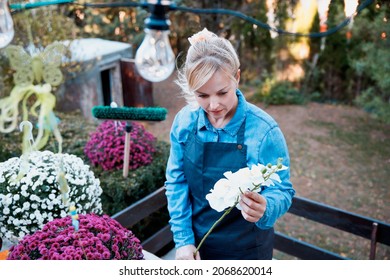 Beautiful Blonde Woman Florist In Her Small Business Outside Her Flower Shop. Female Take Care Of Plants In The Garden Center, Holding White Orchid