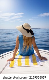 A beautiful, blonde woman with a hat sits on a boat and enjoys the calm sea during her summer holidays