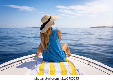 A beautiful, blonde woman with a hat sits on a boat and enjoys the calm sea during her summer holidays