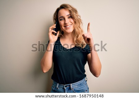 Beautiful blonde woman having conversation talking on the smartphone over white background surprised with an idea or question pointing finger with happy face, number one