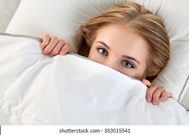 Beautiful Blonde Woman Hiding Face Under Cover Lying In Bed. Female Sparky Eyes Looking In Camera Closeup. Sweet Dreams, Flirtation, Playing Game, Wake Up In Strange Place, Shame, Casual Sex Concept