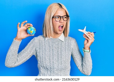 Beautiful Blonde Woman Holding Airplane Toy And World Ball Angry And Mad Screaming Frustrated And Furious, Shouting With Anger. Rage And Aggressive Concept. 