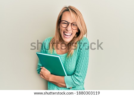 Beautiful blonde woman holding book wearing glasses smiling and laughing hard out loud because funny crazy joke. 