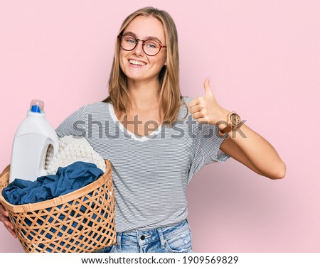 Beautiful blonde woman holding laundry basket and detergent bottle smiling happy and positive, thumb up doing excellent and approval sign 