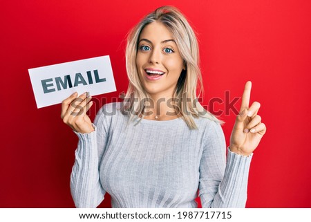 Beautiful blonde woman holding paper with email address smiling with an idea or question pointing finger with happy face, number one 