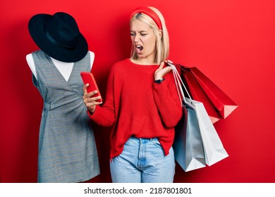 Beautiful Blonde Woman Holding Shopping Bags And Smartphone Angry And Mad Screaming Frustrated And Furious, Shouting With Anger. Rage And Aggressive Concept. 