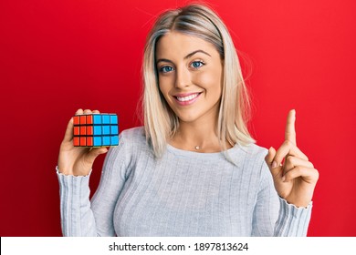 Beautiful Blonde Woman Playing Colorful Puzzle Cube Intelligence Game Smiling With An Idea Or Question Pointing Finger With Happy Face, Number One 