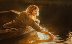 A Beautiful Blonde Woman Sits In An Old Fishing Boat Floating In The Lake And Touches The Water Surface With Her Hand, Illuminated By The Rays Of The Rising Sun. A Journey Into A Fairy Tale.