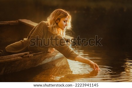 A beautiful blonde woman sits in an old fishing boat floating in the lake and touches the water surface with her hand, illuminated by the rays of the rising sun. A journey into a fairy tale.