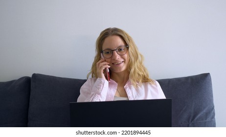 Beautiful Blonde Woman Is Sitting At A Laptop At Home And Talking On The Phone With A Smile. Remote Work From Home Without Visiting The Office. Simplify Work And Reduce Costs.