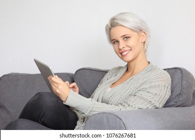 Beautiful blonde woman sitting on a couch and holding a tablet computer in her hand smiling