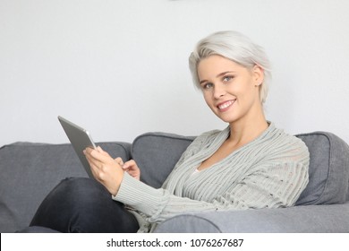 Beautiful blonde woman sitting on a couch and holding a tablet computer in her hand smiling